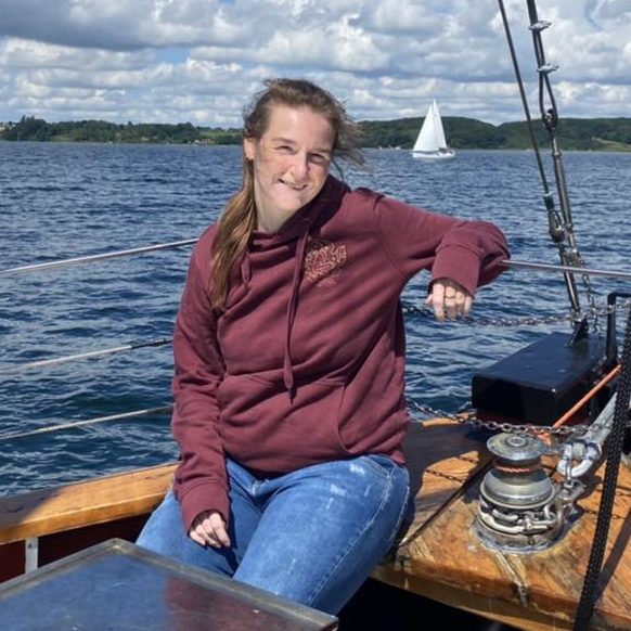 Christa in een bordeauxrode hoodie zit op een zeilboot op rustig water met een zeilboot op de achtergrond.
