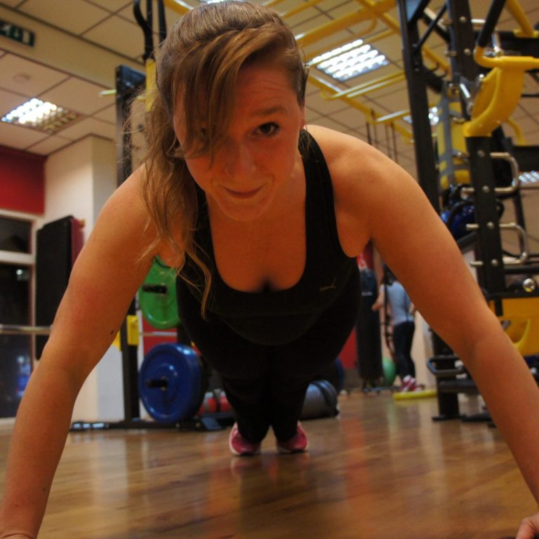 Christa in sportoutfit doet push-ups in een gymzaal met fitnessapparatuur op de achtergrond.