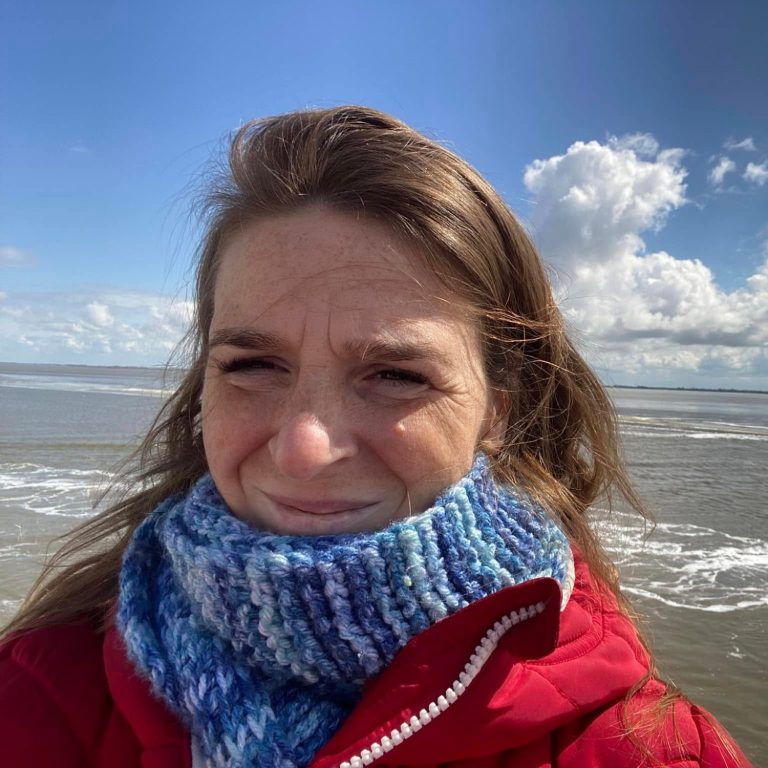 Christa met een blauwe sjaal en rode jas, staande aan zee met wolkenhemel.