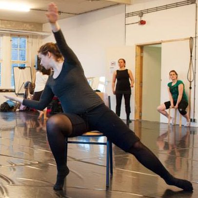 Christa als dansperformer zit op een stoel en reikt naar voren, terwijl anderen toekijken.