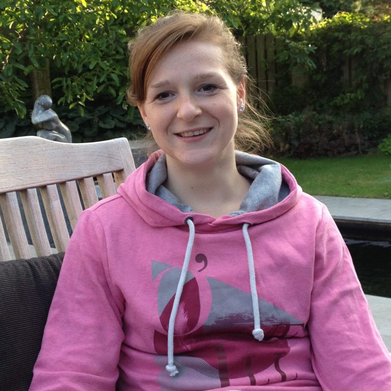 Christa met een roze hoodie, genietend in de tuin met een glimlach.