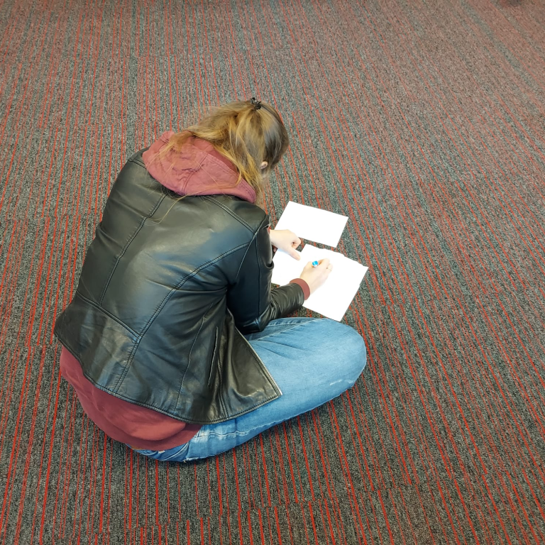 Christa zit op de grond en maakt notities in een schrift, met een pen in de hand.