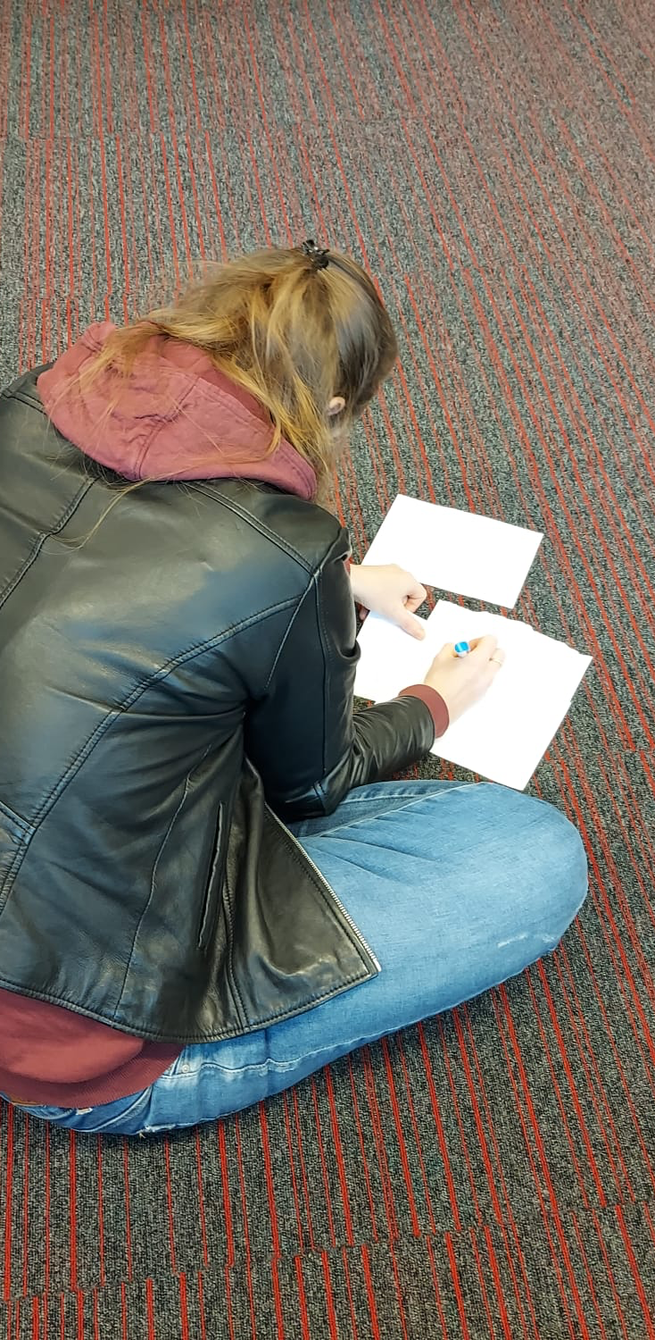 Christa zit op de grond en schrijft op witte blaadjes.