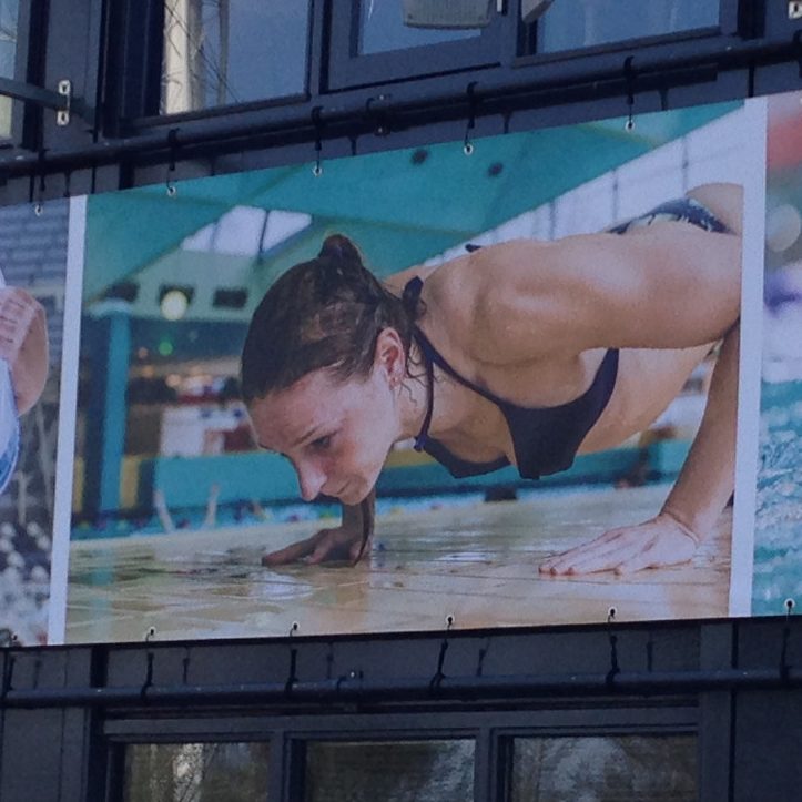 Christa op een billboard doet push-ups aan de rand van een zwembad, in sportoutfit.