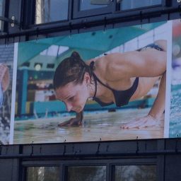 Christa op een billboard doet push-ups aan de rand van een zwembad, in sportoutfit.