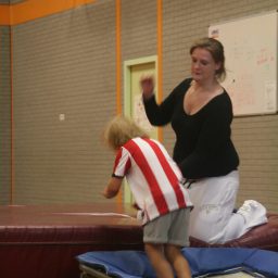Christa helpt een kind bij het springen op een gymmat in een sportzaal.