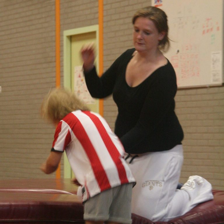 Christa helpt een kind bij het springen op een gymmat in een sportzaal.