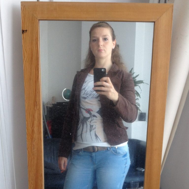 Christa die in een spiegel kijkt, gekleed in een bruine jas en een T-shirt.