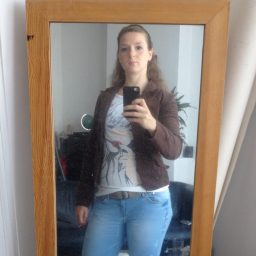 Christa die in een spiegel kijkt, gekleed in een bruine jas en een T-shirt
