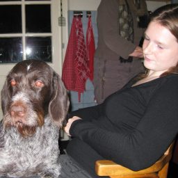 Christa in zwart met een hond op schoot, terwijl een andere persoon op de achtergrond bezig is.