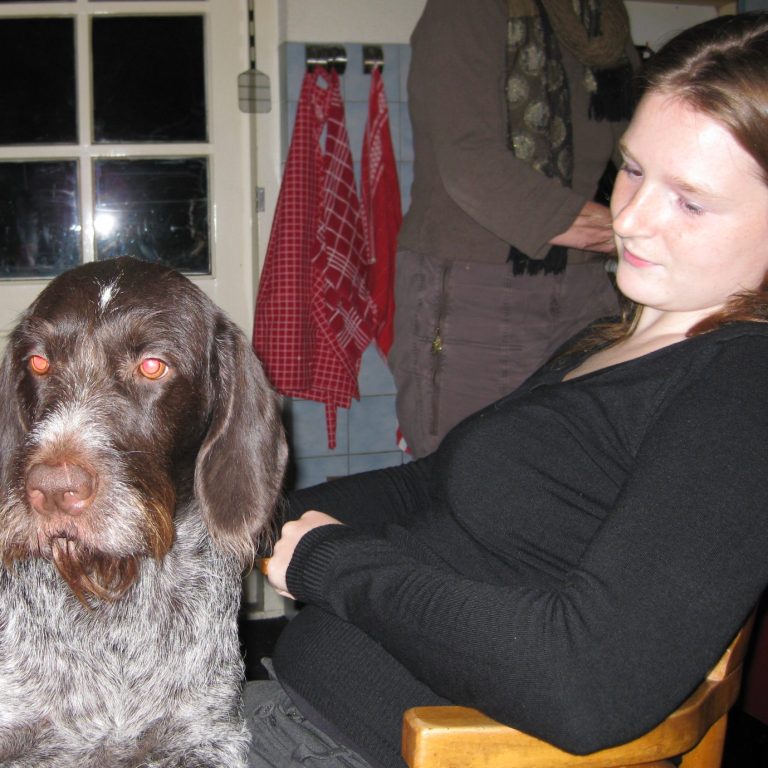 Christa in zwart met een hond op schoot, terwijl een andere persoon op de achtergrond bezig is.