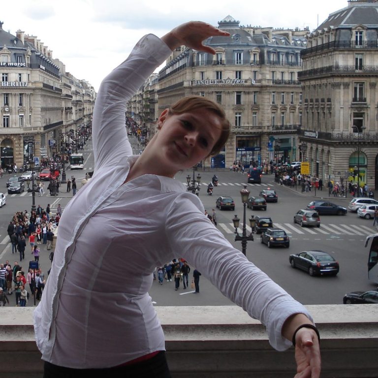 Christa poseert met uitgestrekte armen voor een druk stadsplein.