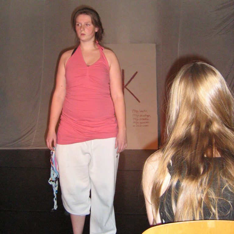 Christa in een roze top en witte broek loopt over een podium tijdens een voorstelling.