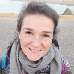 Christa met een glimlach in een casual outfit, zittend buiten met een neutrale achtergrond.